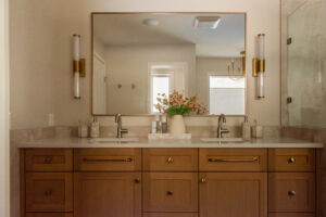 Renovated primary ensuite with warm wood double vanity, brass-framed mirror, wall sconces and glass walk-in shower.