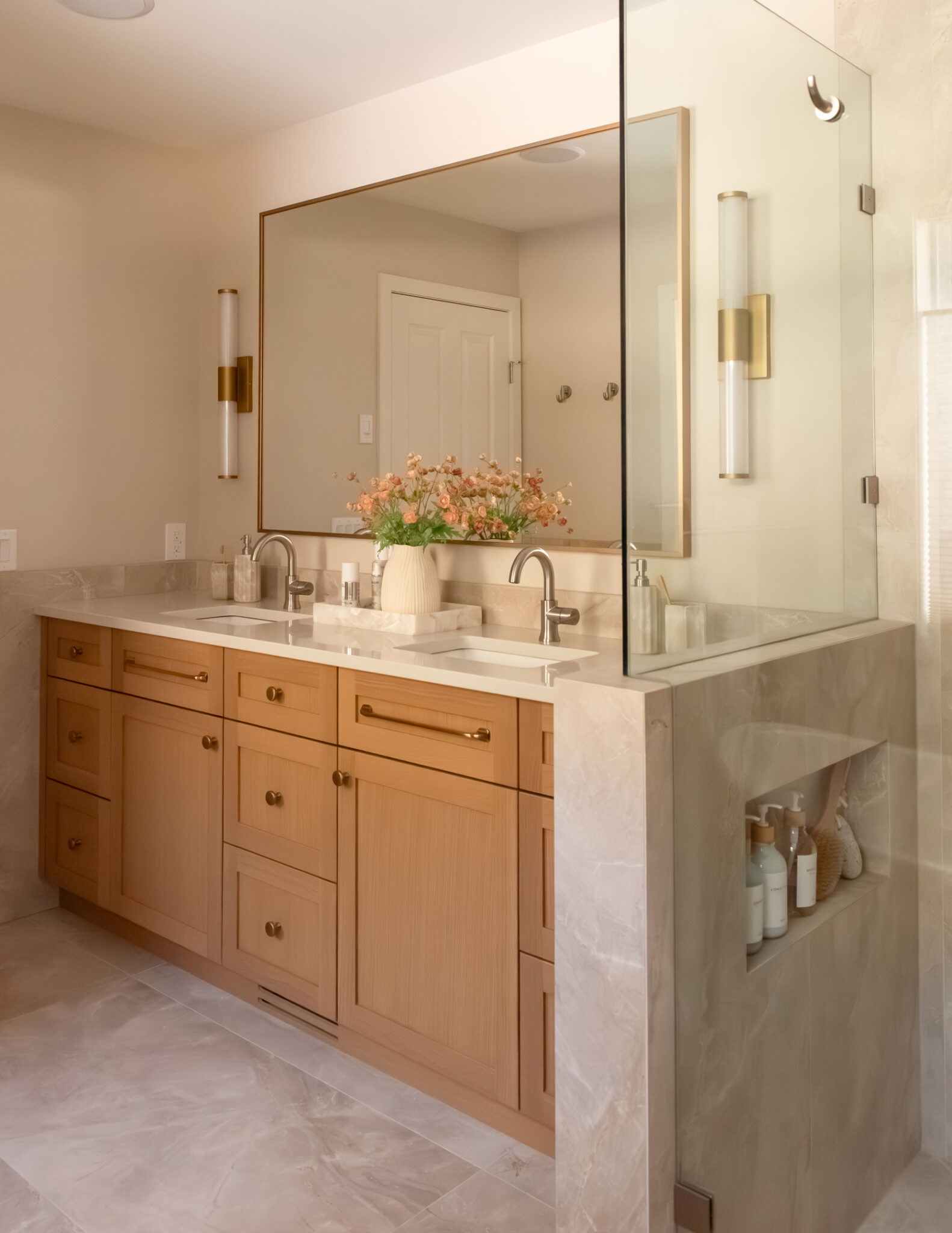 Recessed shower niche built into a stone-look tiled pony wall dividing earthy toned tile shower shower with custom glass and wood toned vanity