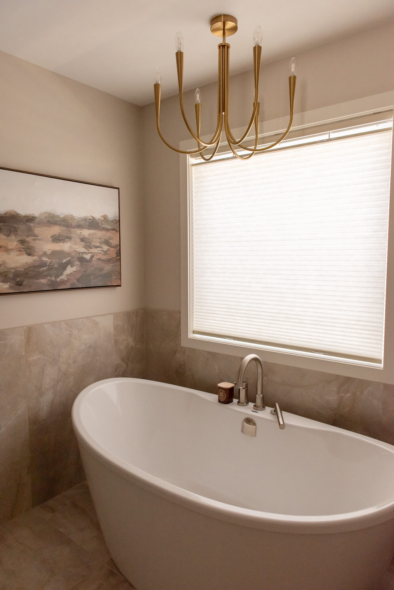 White freestanding soaking tub beneath a window, surrounded by warm stone-look tiled wainscoting and a gold chandelier.