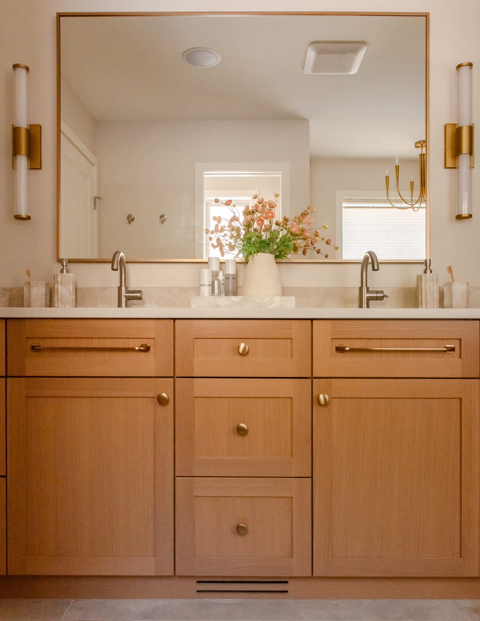 Renovated primary ensuite with warm wood double vanity, brass-framed mirror, wall sconces and glass walk-in shower.