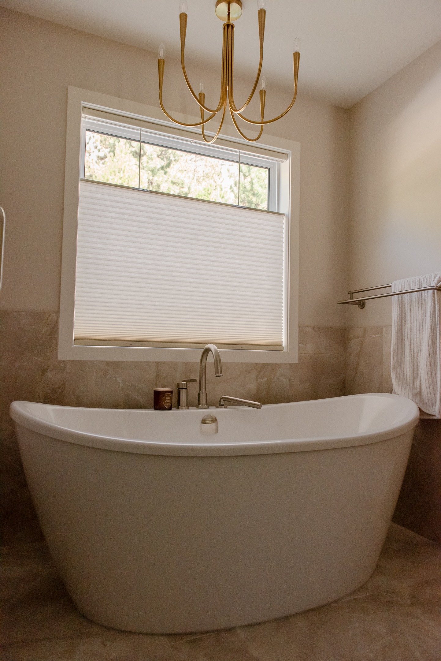 White freestanding soaking tub beneath a window, surrounded by warm stone-look tiled wainscoting and a gold chandelier.