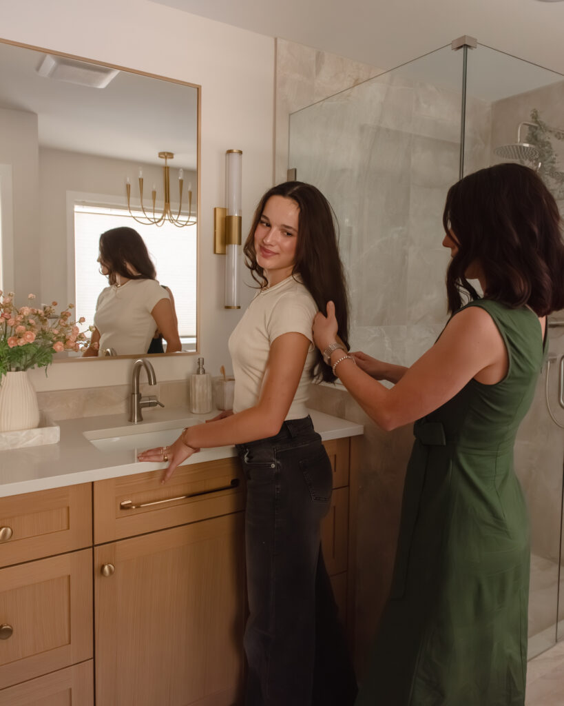 Mother and daughter curling hair in Renovated primary ensuite with warm wood double vanity, brass-framed mirror, wall sconces and glass walk-in shower.