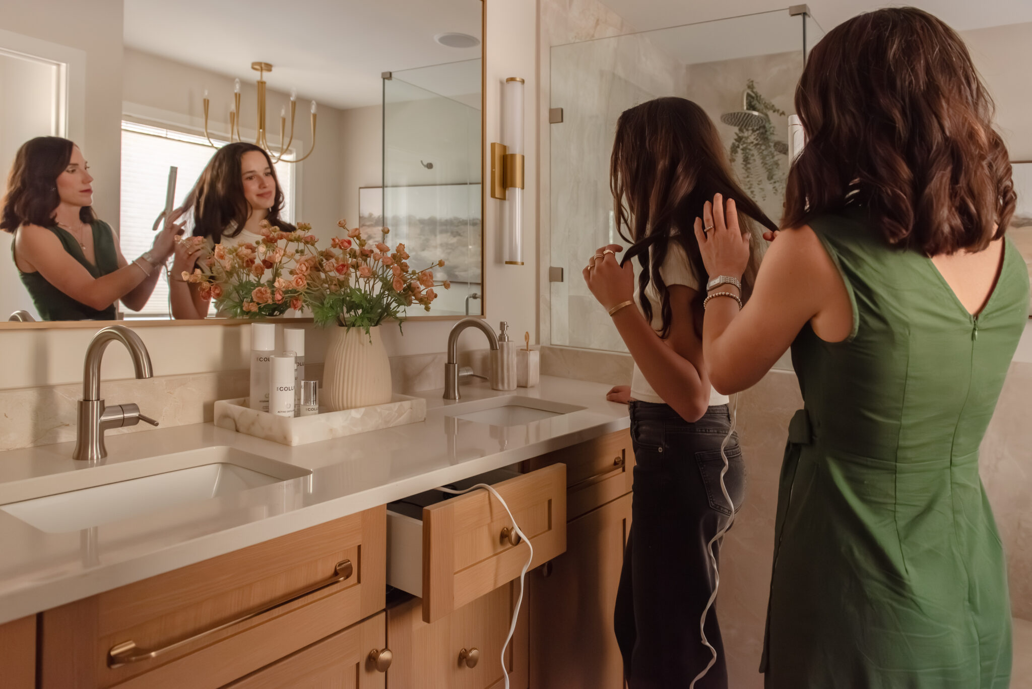 Mother and daughter curling hair in Renovated primary ensuite with warm wood double vanity, brass-framed mirror, wall sconces and glass walk-in shower.