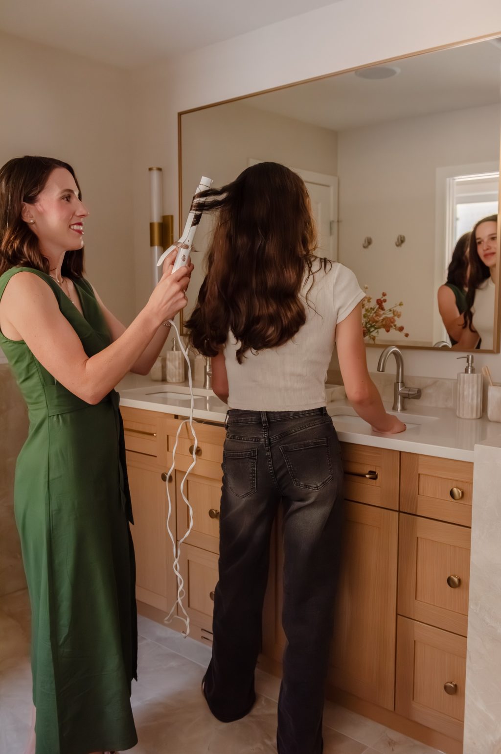 Mother and daughter curling hair in Renovated primary ensuite with warm wood double vanity, brass-framed mirror, wall sconces and glass walk-in shower.